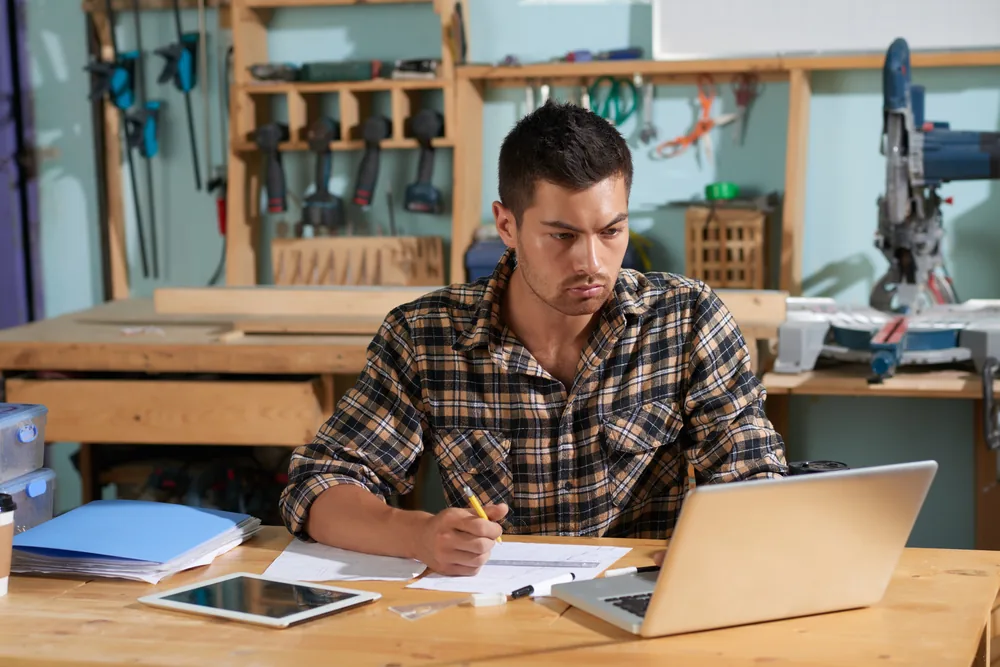 Business owner at a workbench in a shop, going over paperwork and a laptop.