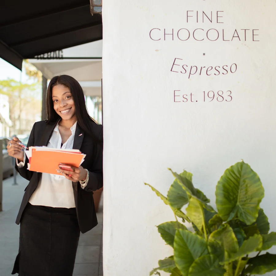 A small business owner standing outside her shop, smiling, with paperwork under her arm